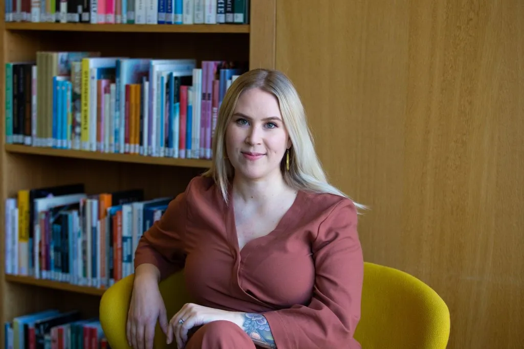 Emma istuu kirjastossa keltaisella tuolilla katsoen hymyillen kameraan.
